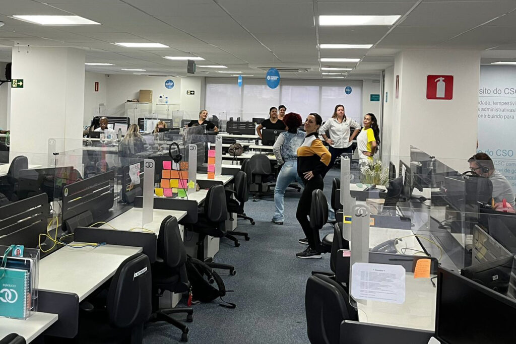 Grupo de colaboradores realizando exercícios de alongamento e ginástica laboral dentro de um escritório com baias de atendimento.
