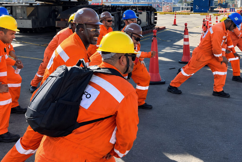 Trabalhadores uniformizados com macacão laranja, capacete de proteção e óculos escuros realizam exercícios de alongamento e ginástica laboral em uma área operacional externa