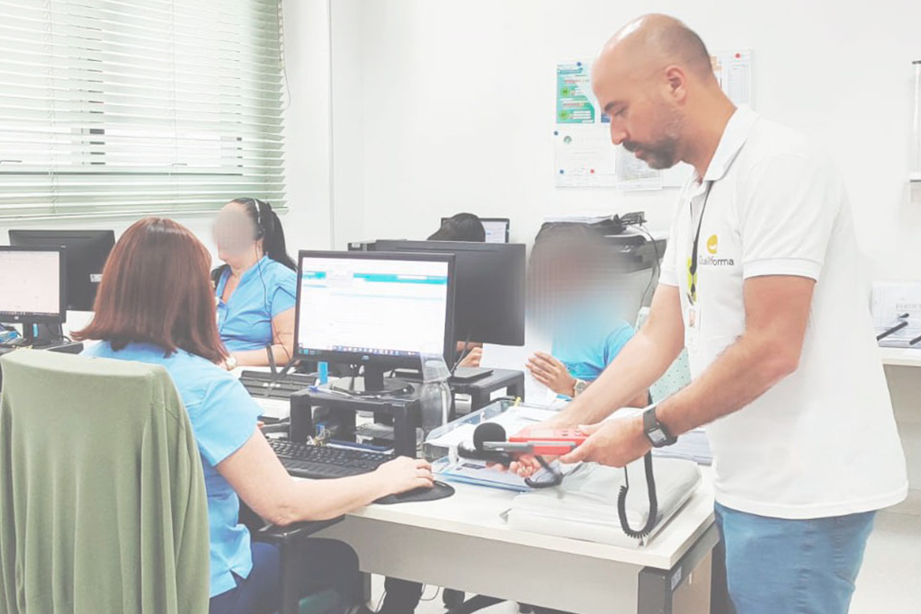 Técnico da Qualiforma vestindo camisa polo branca com logotipo mede o nível de ruído com um equipamento de medição digital em um escritório de call center enquanto colaboradoras de uniforme azul trabalham nos computadores.