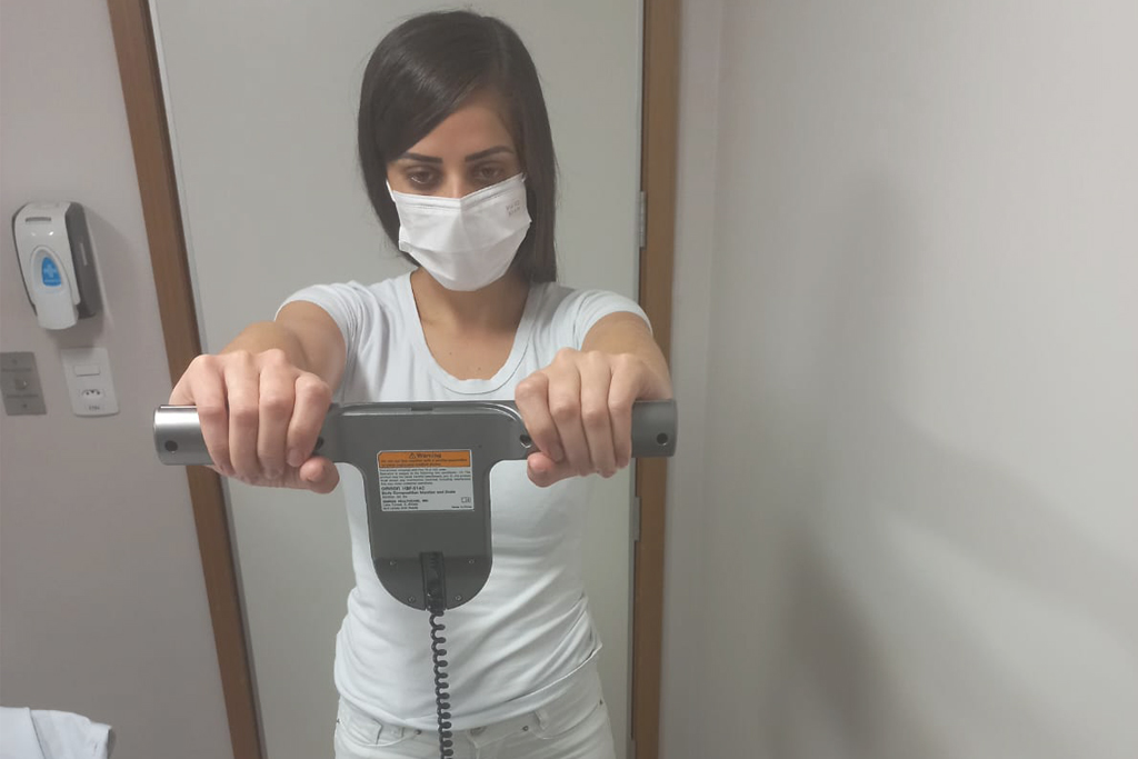 Profissional de saúde da Qualiforma usando máscara de proteção e camiseta branca segura as manoplas superiores de uma balança de bioimpedância para exame de composição corporal.