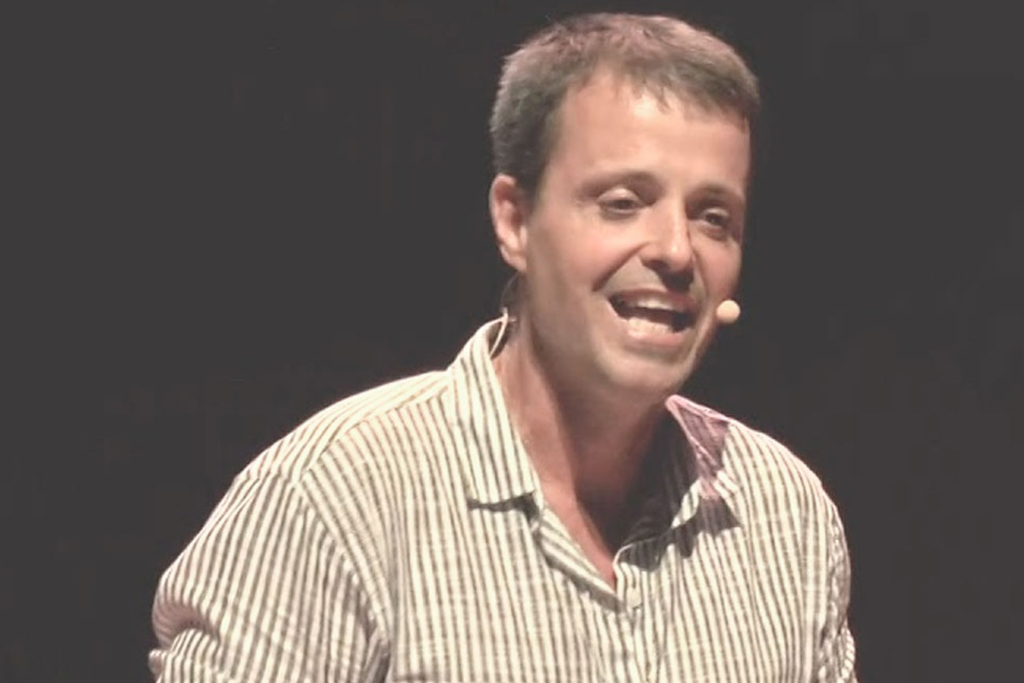 • Homem palestrante sorrindo com microfone auricular fala no palco durante um evento corporativo com fundo escuro.