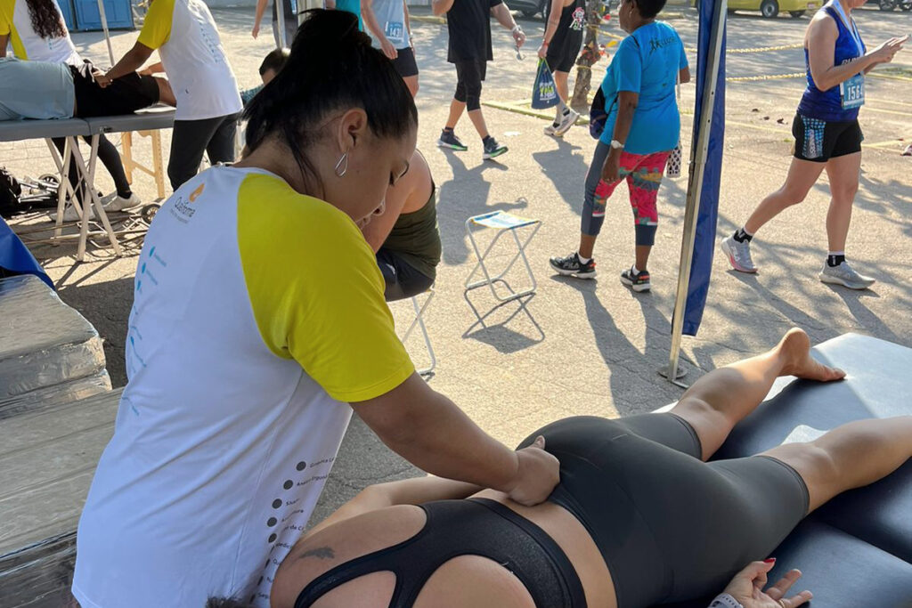 Profissional da Qualiforma vestindo camiseta branca e amarela realiza massagem relaxante nas costas de uma atleta deitada em uma maca durante uma etapa do circuito das estações