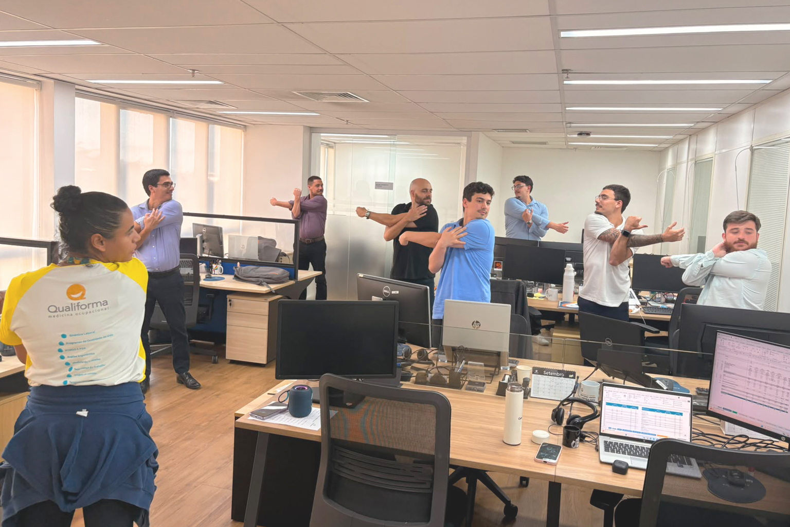 Instrutora da Qualiforma vestindo uniforme branco e amarelo orienta um grupo de colaboradores que realiza alongamento de braço em pé nas baias de um escritório.